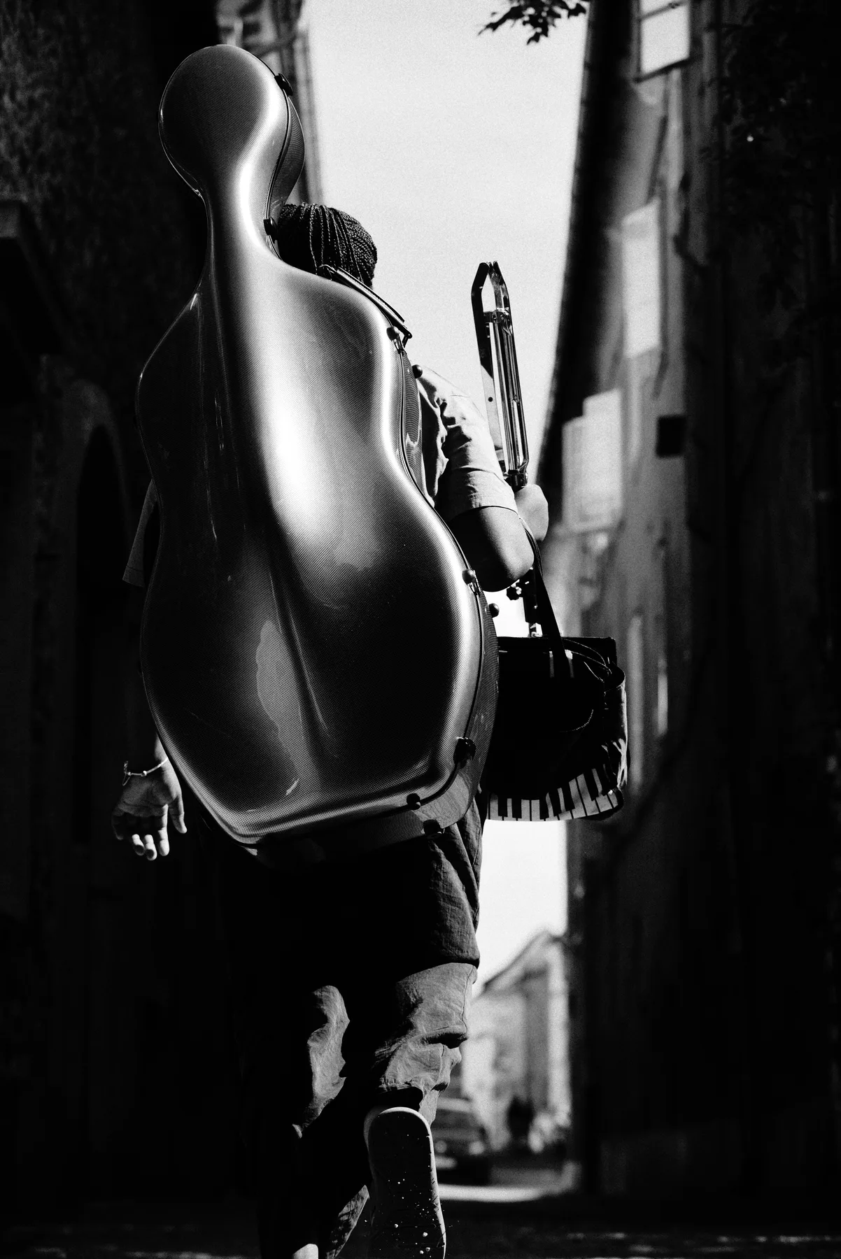 A black-and-white close-up of a musician carrying an instrument case along a narrow street.