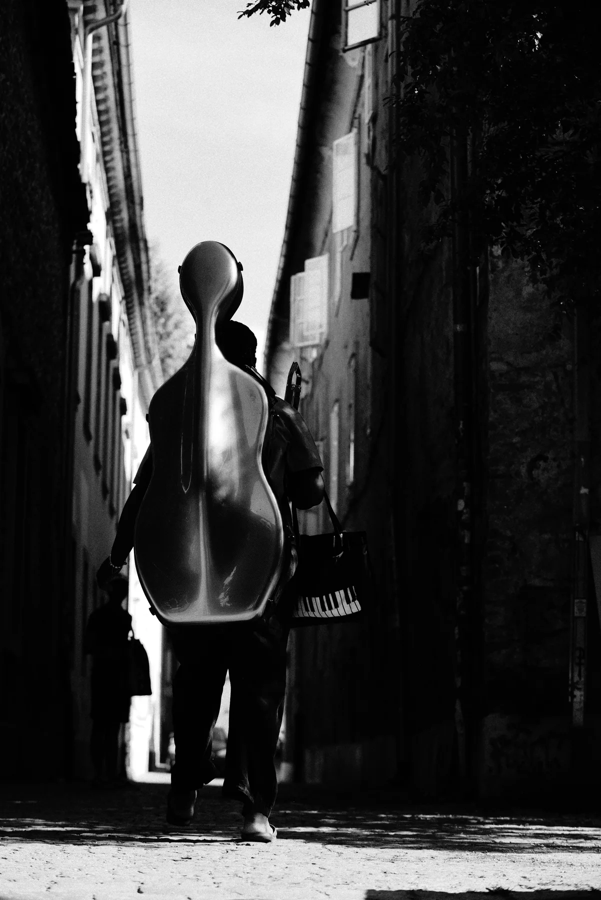A black-and-white view of a musician carrying an instrument case through a narrow street.
